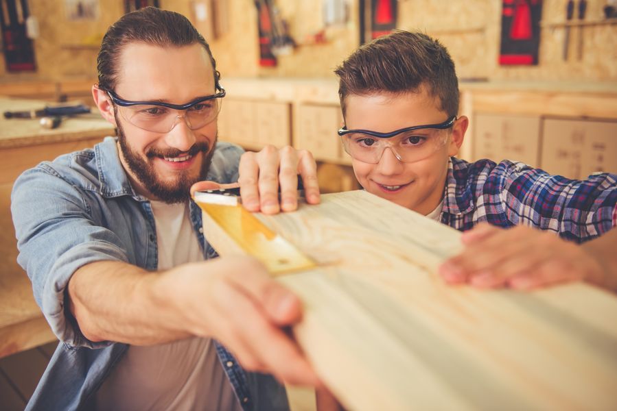 Handsome teenage carpenter and his father in protective glasses are measuring wood and smiling while working in the workshop
